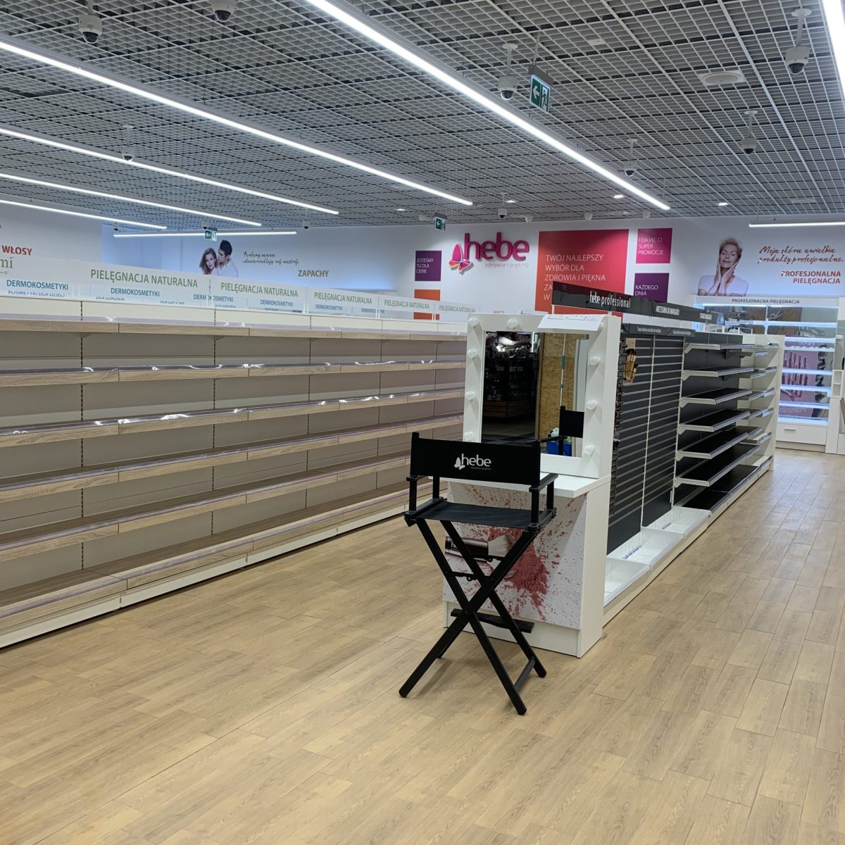 Wnętrze jasnego sklepu (prawdopodobnie drogerii) z pustymi lub częściowo pustymi półkami. Podłoga wyłożona jest jasnymi panelami imitującymi drewno. W centrum kadru stoi czarne, składane krzesło w stylu reżyserskim z napisem na oparciu. Na ścianach w tle widoczne są reklamy.