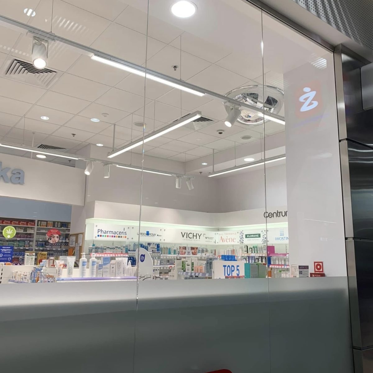 Widok z zewnątrz na witrynę nowoczesnej apteki. Za dużą szklaną szybą widoczne są białe wnętrza, oświetlone lampami LED. Na półkach znajduje się wiele produktów farmaceutycznych i kosmetycznych, w tym widoczne logo VICHY.