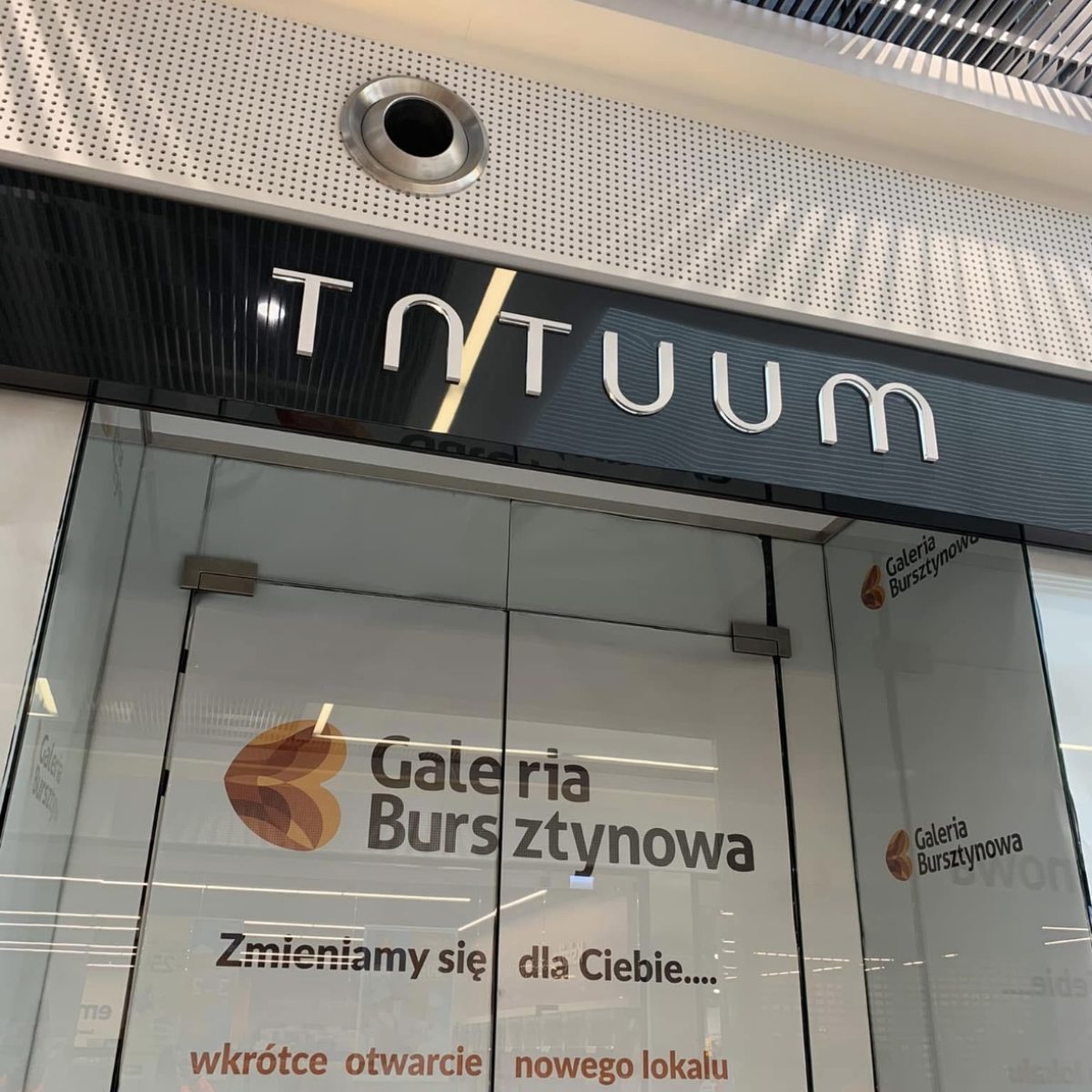 Zdjęcie wejścia do sklepu z napisem **"TATUM"** nad drzwiami. Na szklanych drzwiach widnieje logo i nazwa **"Galeria Bursztynowa"** oraz komunikat: "Zmieniamy się dla Ciebie.... wkrótce otwarcie nowego lokalu". Powyżej wejścia widoczne są elementy wentylacji i perforowany sufit centrum handlowego.