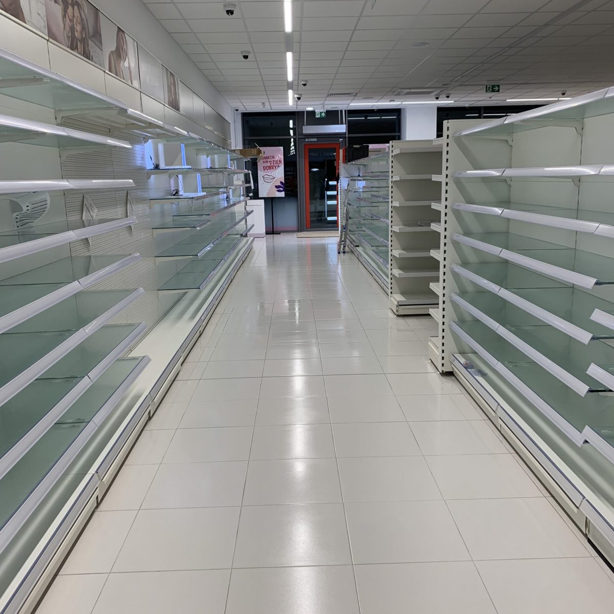 Długie, puste przejście w sklepie, z rzędami białych regałów z każdej strony. Podłoga jest wyłożona białymi, lśniącymi płytkami. W tle widoczne jest wejście do innego pomieszczenia lub strefy kas, oświetlone na czerwono.