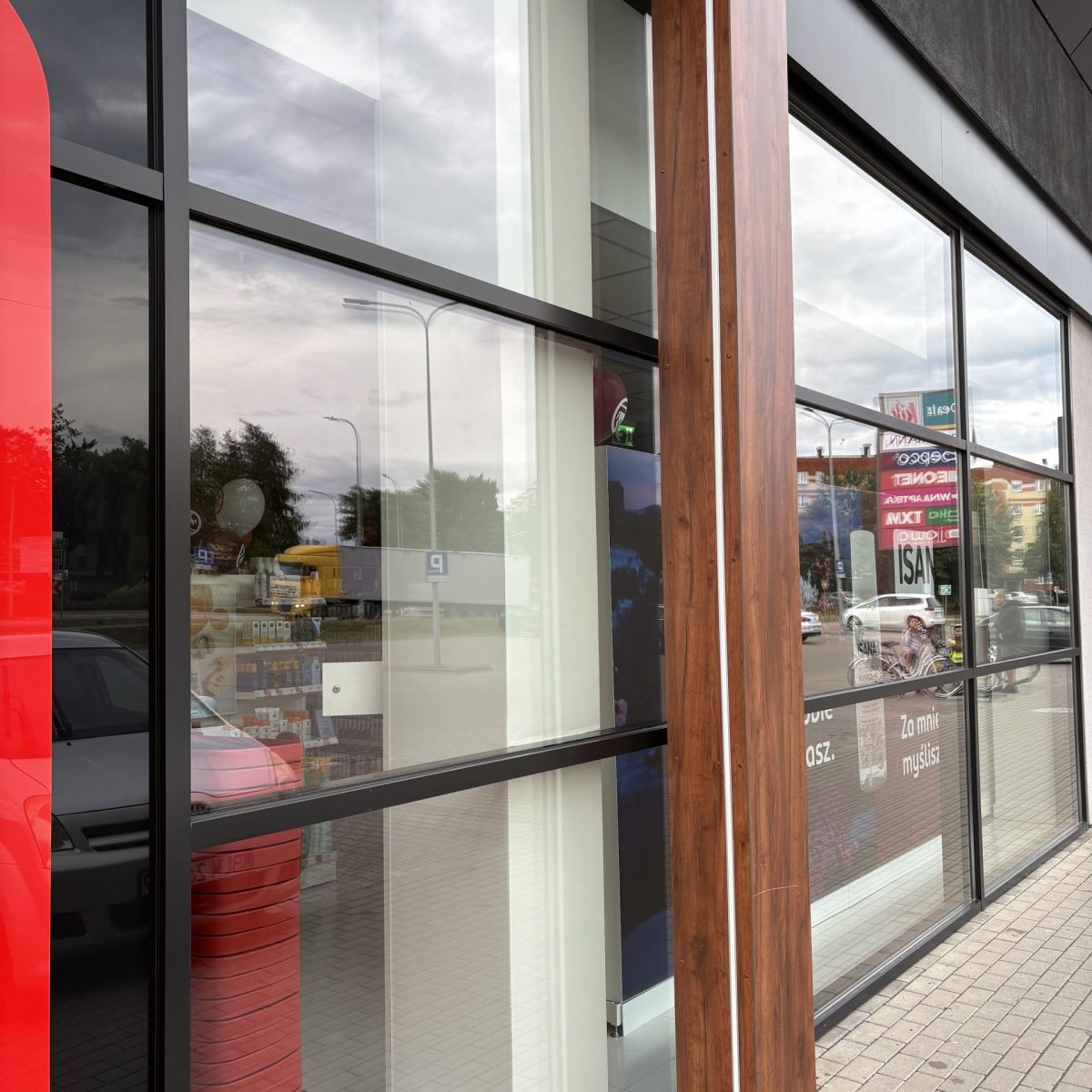 Zewnętrzny widok na nowoczesną, przeszkloną witrynę sklepową. Duże szyby są rozdzielone ciemnymi, metalowymi ramami i jednym pionowym słupem obłożonym drewnopodobnym materiałem. W odbiciu widać pochmurne niebo, a przez szybę prześwituje wnętrze i fragment ulicy.