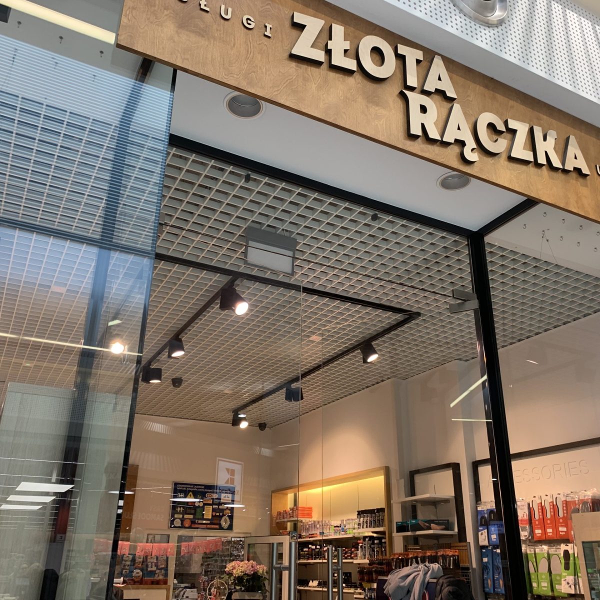 Zdjęcie witryny i wejścia do punktu usługowego z drewnianym szyldem z napisem **"ZŁOTA RĄCZKA"** (częściowo zasłonięty) oraz **"USŁUGI"**. Wnętrze jest oświetlone reflektorami na czarnych szynach i widoczne są w nim półki z różnymi przedmiotami. Sklep znajduje się w centrum handlowym z kratownicowym sufitem.