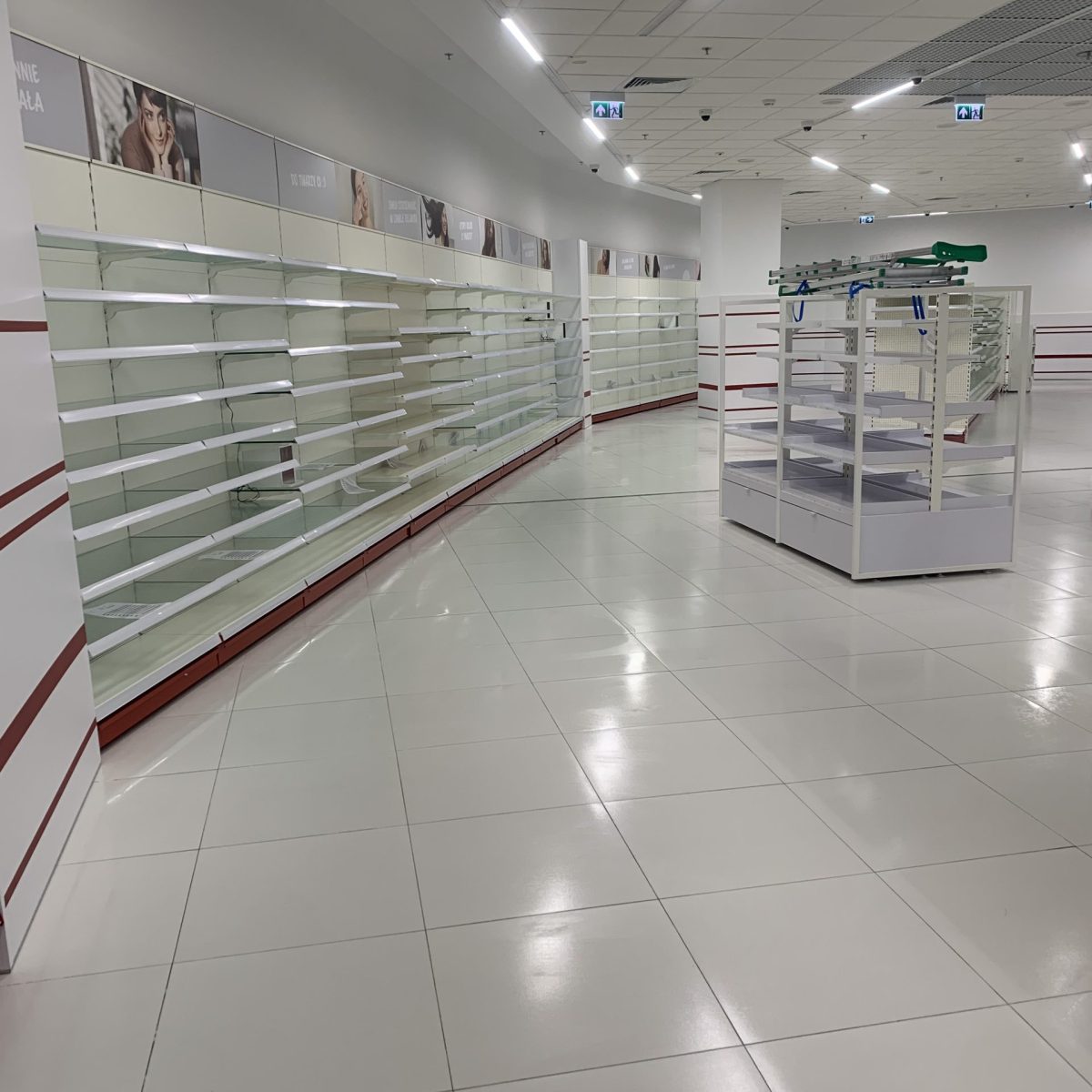 Szeroki widok na wnętrze dużego, pustego sklepu o białym wykończeniu. Podłoga wyłożona jest dużymi, białymi płytkami. Wzdłuż ścian ciągną się długie rzędy pustych półek sklepowych z czerwonymi akcentami. W centrum kadru widoczny jest wolnostojący regał ekspozycyjny.